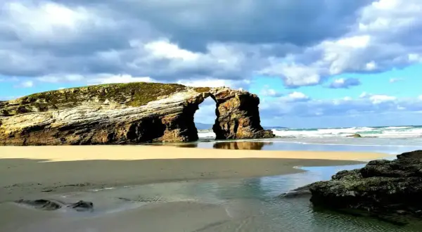 Arco natural en las playas de Galicia, con arena dorada, mar azul y cielo nublado.
