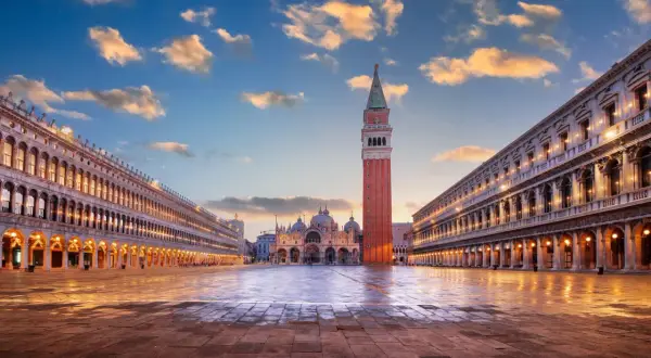 La piazza San Marco s'éveille sous un ciel aux teintes orangées. La basilique et le campanile se dressent majestueusement, reflétés sur le sol mouillé.