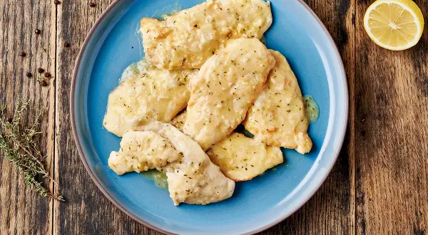 Piatto di pollo al limone su un piatto blu, con limone e rosmarino sullo sfondo.