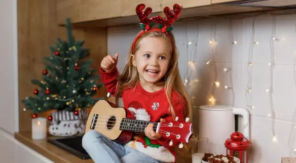 Dziewczynka w świątecznych rogach renifera gra na ukulele w kuchni, obok choinki i lampek.