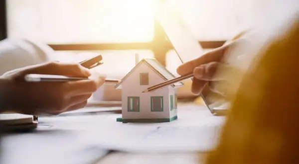 Mani che studiano un modello di casa, con matite e calcolatrice. Si discute il surroga mutuo significato per un nuovo acquisto immobiliare.