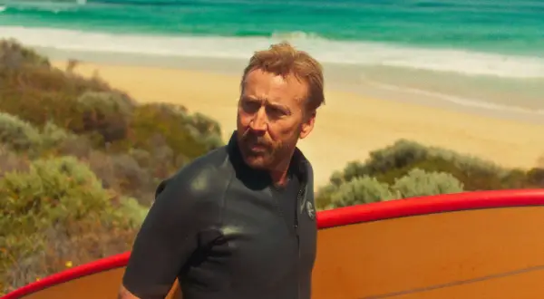 Nicolas Cage w piance z deską surfingową na plaży, jak w jednym z jego filmów.