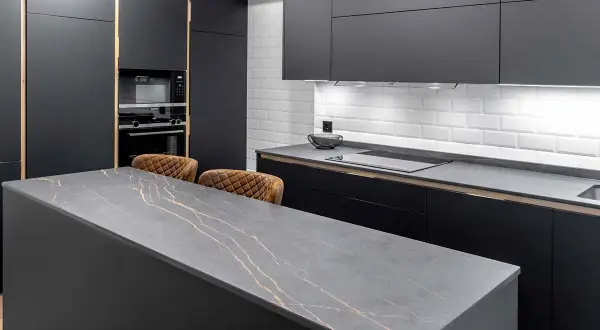 Isla de cocina moderna con encimera de piedra gris veteada en oro. Dos taburetes de cuero marrón esperan junto a la medida encimera.