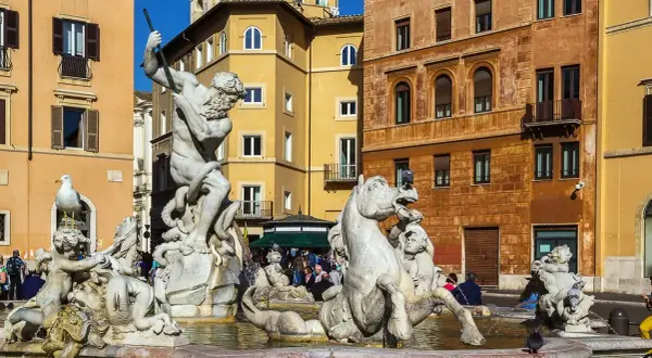 La Fuente de Neptuno en Roma, con su imponente estatua de mármol luchando contra un pulpo, rodeada de caballos marinos y figuras mitológicas, bajo el sol y edificios coloridos.