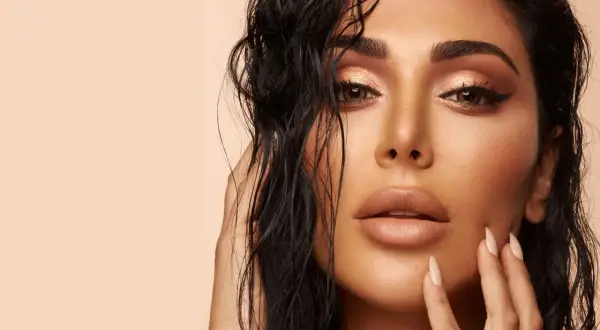 Un visage de femme au maquillage sophistiqué, avec des yeux cuivrés et des lèvres en teinte nude. Ses cheveux mouillés encadrent son visage.