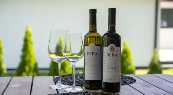 Dwa kieliszki i dwie butelki wina Bella Classy, białe i czerwone, bezalkoholowe, na drewnianym stole w ogrodzie.
