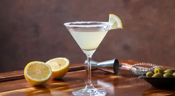 Elegancki martini bianco drink z solnym rantem i plasterkiem cytryny, obok połówki cytryny, oliwki i akcesoria barmańskie.