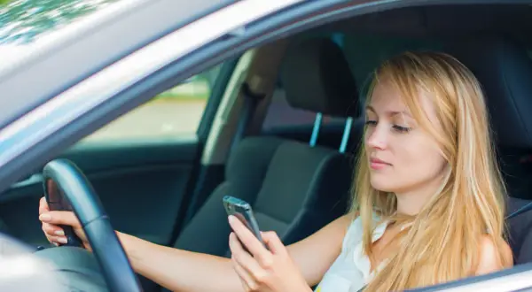 Mujer rubia al volante, mirando su móvil. Al distraerse el conductor tarda, poniendo en riesgo su seguridad y la de otros.