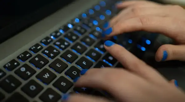 Dłonie z niebieskim lakierem na paznokciach piszą na podświetlanej klawiaturze MacBooka. Twarda spacja jest widoczna.