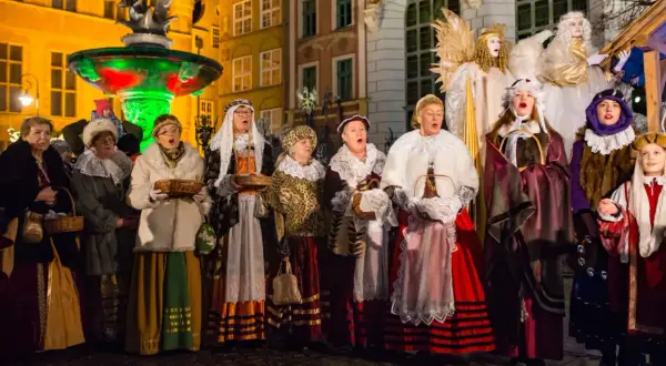 Grupa kobiet w strojach historycznych śpiewa kolędy. W tle iluminowana fontanna i zabytkowe kamienice.