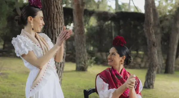 Dos mujeres vestidas para ir al Rocío, con trajes blancos y mantones, sonríen y aplauden en un jardín.
