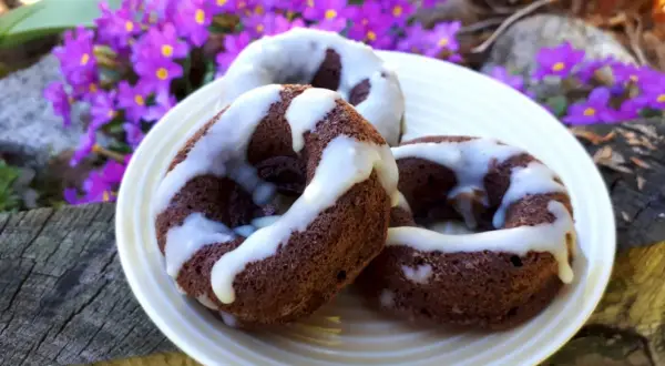 Trzy czekoladowe donuty brownie polane białym lukrem, na talerzu wśród fioletowych kwiatów.