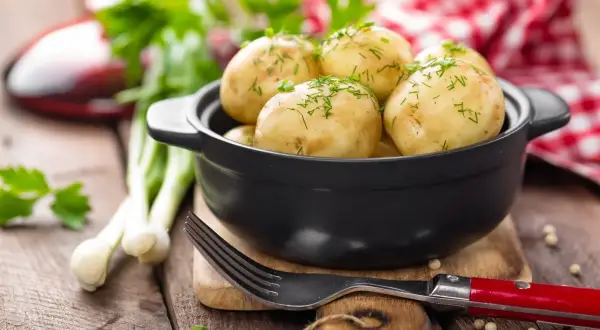 Młode ziemniaki w czarnej misce, posypane koperkiem, obok widelca i dymki.