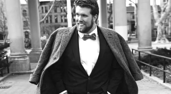 Un homme gros bien habillé, souriant, porte un costume sombre, une chemise blanche et un nœud papillon. Son manteau est jeté sur ses épaules.