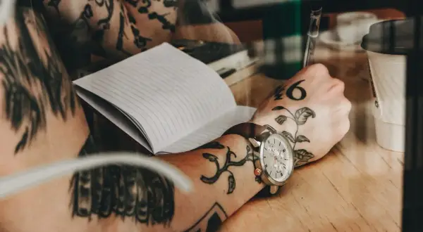 Dłoń z tatuażem na mostku pisze w notatniku. Na nadgarstku zegarek.