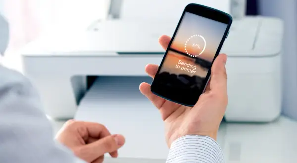 Dłoń trzyma smartfon z napisem "Sending to printer", pokazując jak drukować z telefonu. W tle biała drukarka.