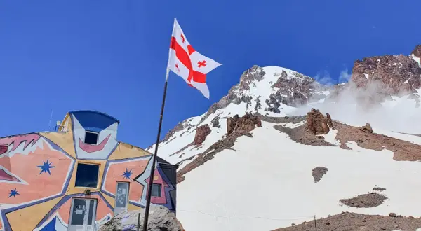 Kolorowy budynek i gruzińska flaga na tle ośnieżonych szczytów. Kazbek wysokość robi wrażenie.