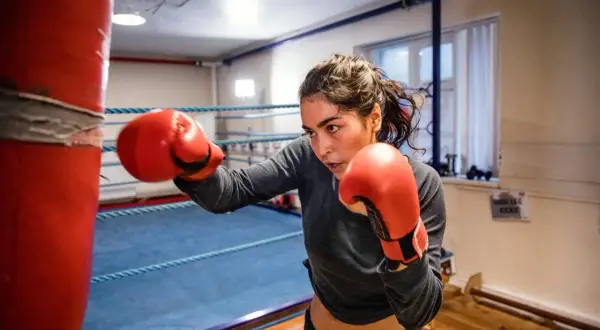 A determined woman in red boxing gloves practices her boxing jabbing technique on a red punching bag in a boxing ring.