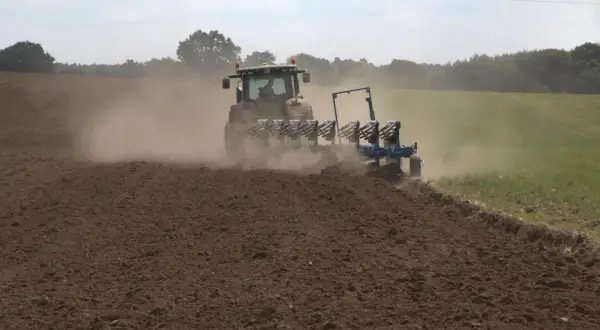 Traktor z pługiem orze pole, wzbijając tumany kurzu. To przygotowanie gleby do kolejnego sezonu, być może z myślą o wapnowaniu gleby.