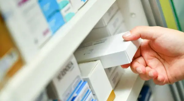 Dłoń sięga po białe pudełko leku, być może zawierającego nilogrin, z aptecznej półki pełnej innych opakowań.