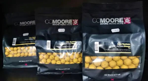 Trzy opakowania kulek proteinowych CC Moore Live System w różnych rozmiarach. Kulki są żółte, widoczne przez przezroczyste okienko.