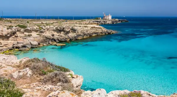 Scogliera rocciosa con mare cristallino e un faro in lontananza, un angolo di paradiso tra le favignana spiagge.