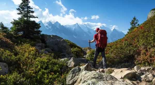 Randonneur avec sac à dos rouge, bâtons de marche, sur un sentier rocheux. Montagnes enneigées en arrière-plan. Combien de jours de marche pour atteindre ce sommet ? gr5.
