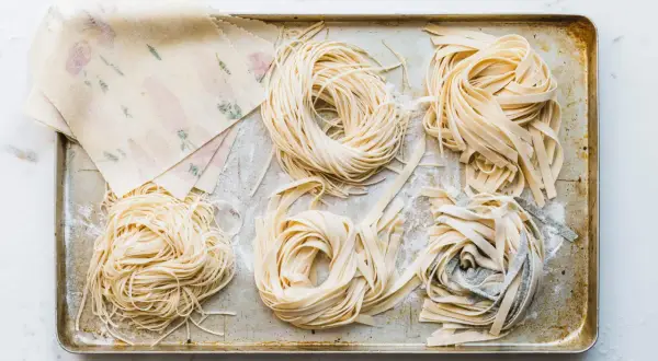 Frische Nudeln in verschiedenen Formen liegen auf einem Blech. Ein Stapel mit Blumenmuster verzierter Teigblätter liegt daneben. Perfekt für alle, die Nudeln selber machen wollen.