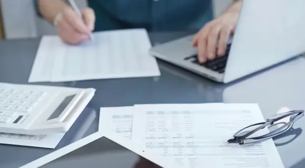 Osoba pracuje przy laptopie, przeglądając dokumenty i pisząc. Na biurku leży kalkulator i okulary. Przygotowania do podpisania umowy zlecenie 2026.