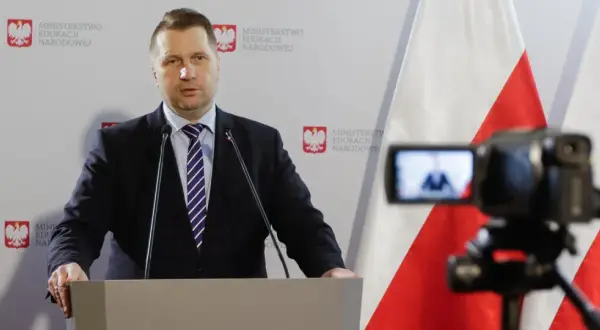 Mężczyzna w garniturze przemawia przy mównicy na tle flagi Polski i logo Ministerstwa Edukacji Narodowej.