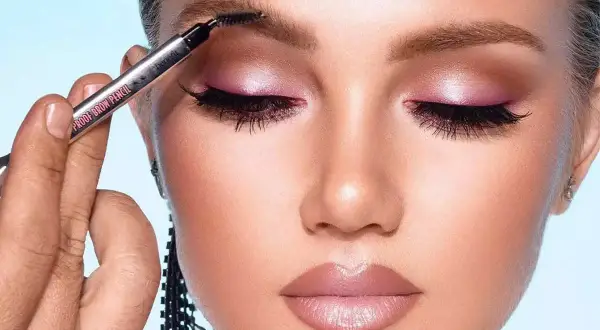 Occhi con ombretto rosa scintillante e labbra morbide. Una mano sfuma le sopracciglia con un pennellino, completando un look da sogno.