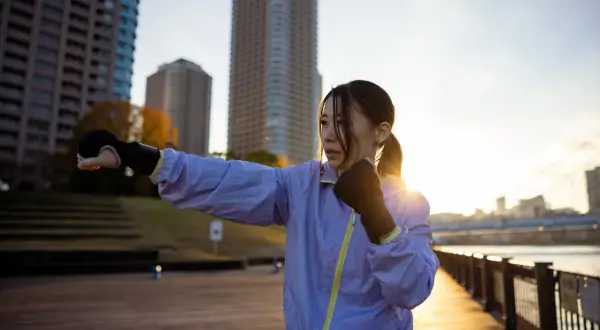 A woman practices shadow boxing outdoors, a popular fitness trend, with city buildings in the background at sunset.