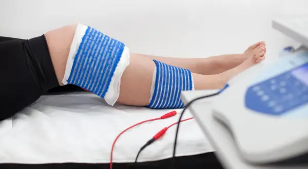 Elektrody podłączone do urządzenia rehabilitacyjnego przy nodze, gdzie widoczna jest kość piszczelowa.