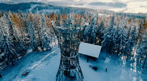 Wieża widokowa na borowej górze w zimowej scenerii. Śnieg pokrywa drzewa i ziemię, a ludzie spacerują wokół konstrukcji.