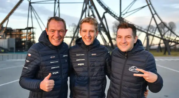 Trzech mężczyzn w kurtkach z logo Energylandii pozuje na tle konstrukcji roller coastera.