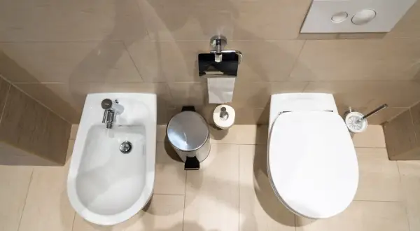 Łazienka z bidetem i WC. Widać kosz na śmieci, papier toaletowy i szczotkę. Odległość WC od ściany bocznej jest optymalna.