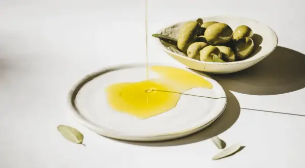Flüssiges Olea europaea fruit oil fließt auf einen Teller, daneben eine Schale mit grünen Oliven und Blättern.