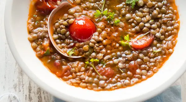 Una ciotola bianca piena di saporita zuppa di lenticchie con pomodorini e prezzemolo fresco. Un mestolo serve la zuppa.