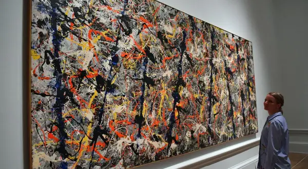 A woman admires a massive Jackson Pollock painting worth $140 million, a chaotic symphony of splattered paint in black, white, orange, and blue.