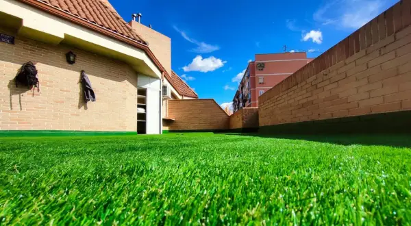 Un vibrante césped artificial para parques infantiles cubre una terraza con edificios de ladrillo y un cielo azul brillante.