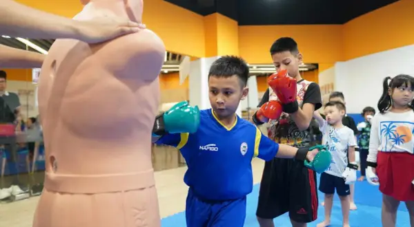 Young boxers practice their moves, with one boy demonstrating boxing blocking techniques on a training dummy.