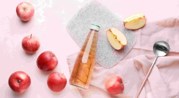 Frische Äpfel und eine Flasche mit Apfelessig mit Wasser. Ein Löffel liegt daneben.