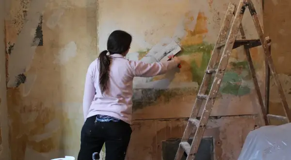 Kobieta z pędzlem w ręku usuwa tynk strukturalny ze ściany. Obok stoi drabina.