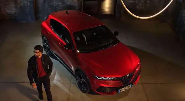 Elegancki mężczyzna w okularach obok lśniącego, czerwonego Alfa Romeo Tonale. Stylowy duet w industrialnym wnętrzu.