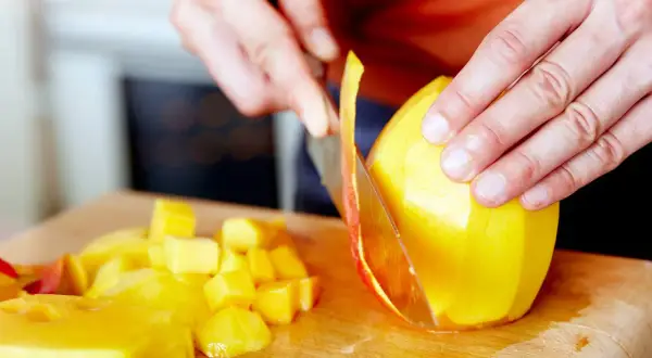 Hände schneiden eine reife Mango auf einem Holzbrett. Saftige Fruchtstücke liegen daneben.