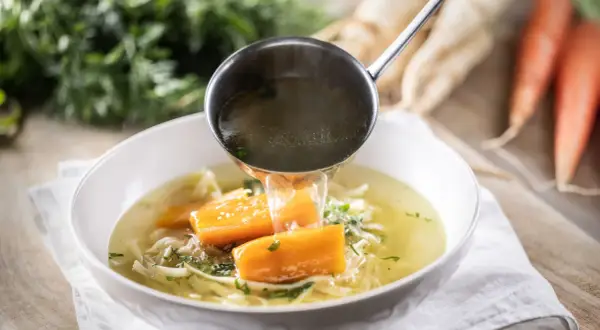 Un mestolo versa brodo caldo in una ciotola di zuppa di pollo con carote e tagliatelle.