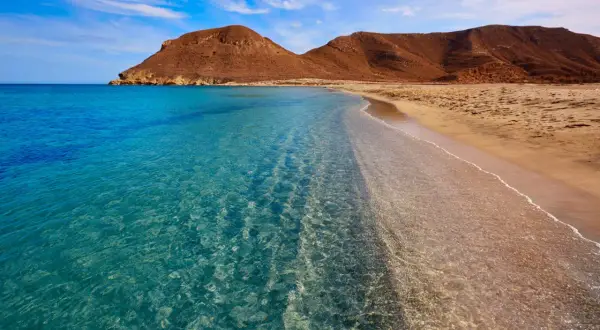 Aguas cristalinas acarician la arena dorada de las playas vírgenes de Almería, con montañas áridas de fondo bajo un cielo azul.