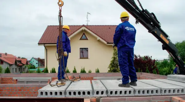 Dwóch budowlańców w żółtych kaskach montuje prefabrykowane płyty stropowe.