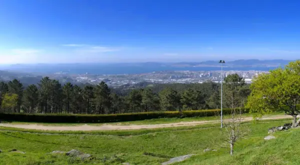 Vista panorámica desde el monte dos pozos, con un camino de tierra y césped en primer plano, árboles y un faro, y una ciudad y el mar al fondo.