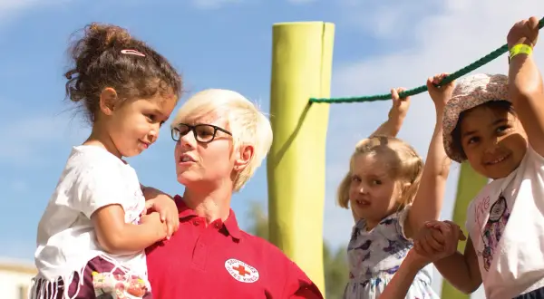 Frau vom Roten Kreuz fördert **Inklusion im Kindergarten**, indem sie ein Kind hält, während andere Kinder an einem Seil spielen.